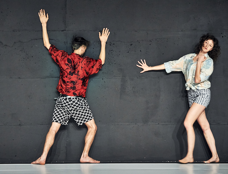 Two dancers in geometric-printed shorts and tropical button ups lean against a concrete wall for support.